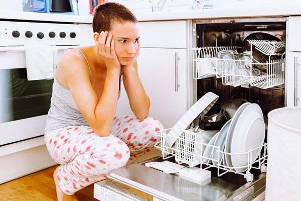 teenage girl with short hair, in home clothes, clutched head from fatigue from home routine. filled dishwasher with dirty dishes in home kitchen is not functioning, it has broken.