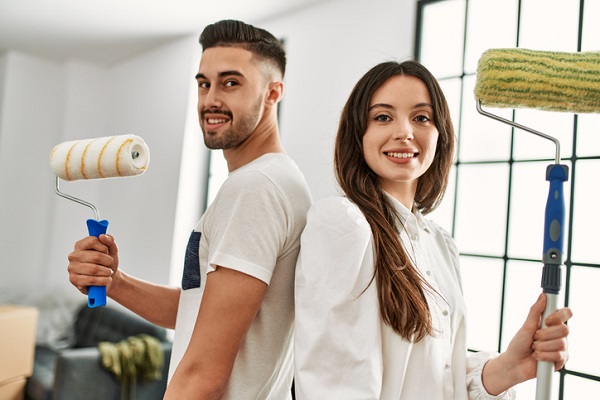 Young couple smiling and looking at each other while holding painting tools, taking a break from painting the walls in their new home